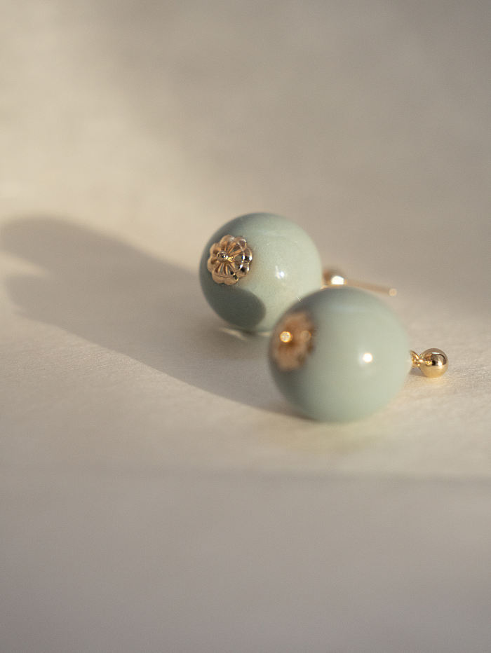 Close-up of celadon porcelain earrings in warm morning light, showcasing crackled glaze and soft green hue.

