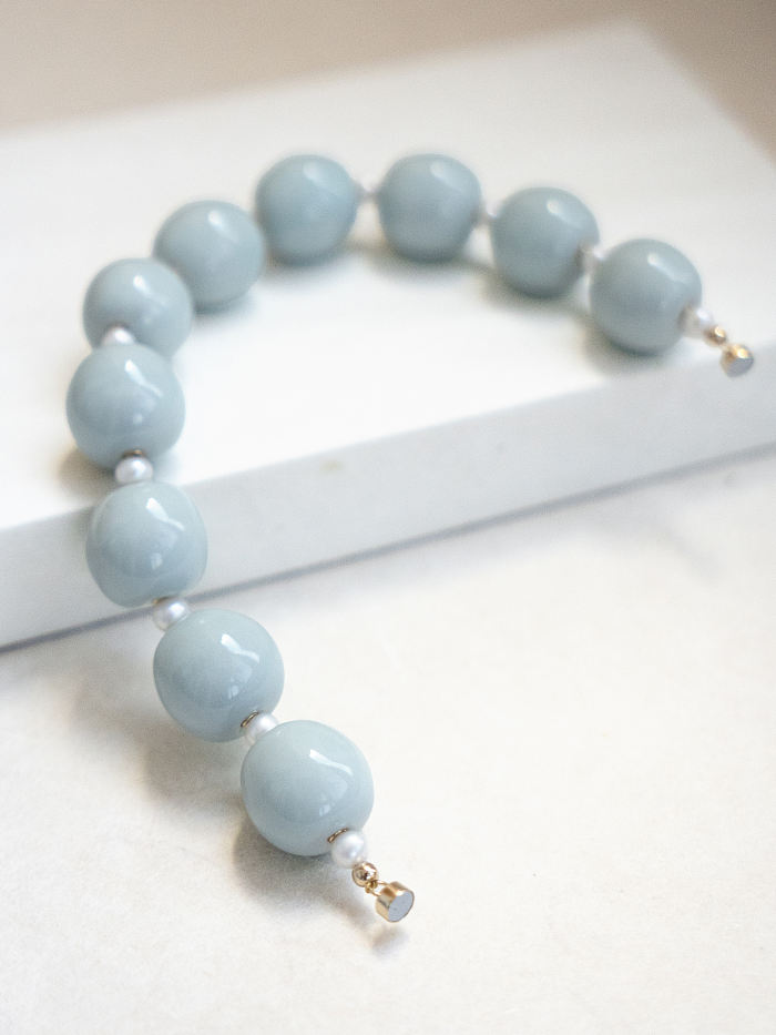 Soft-lit flatlay of the bracelet partially draped over a white block, revealing the gold magnetic clasp and glaze reflections.