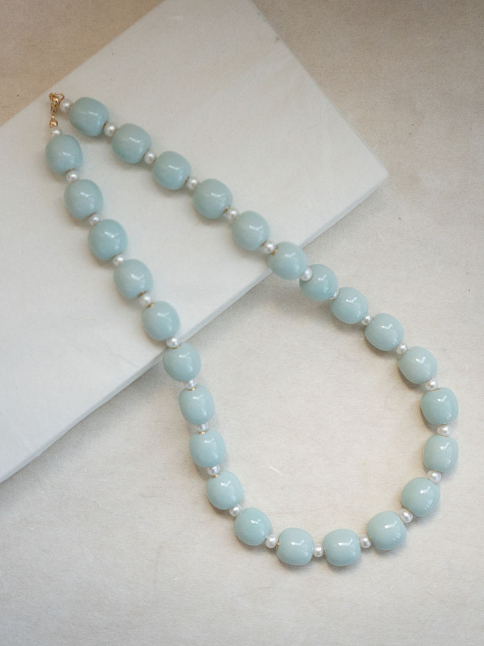 Flatlay of the necklace gently curved on a white book, highlighting bead rhythm and pale blue-green tones.