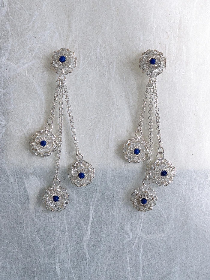 Top-down view of Huasi Cascade Earrings on white and grey paper background, showing soft chain drape and floral spacing in static composition